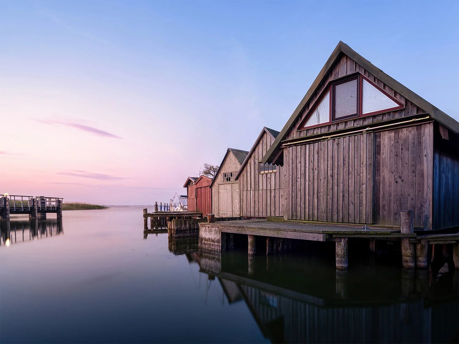 Hölzerne Bootshäuser stehen auf Stegen im stillen Wasser, darüber ein rosa-blauer Abendhimmel